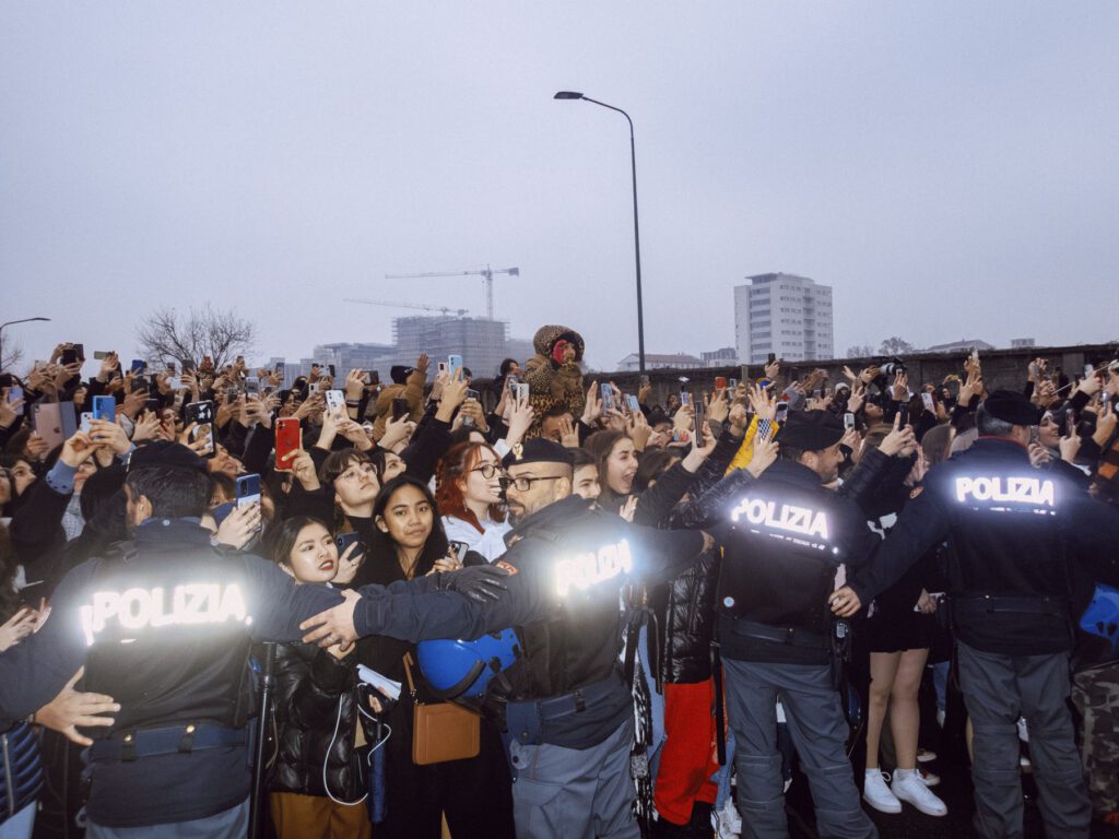 A crowd of people is captured holding up smartphones to take pictures or record an event. Four police officers with "POLIZIA" on their jackets are seen facing the crowd, seemingly there to maintain order. The crowd is diverse, with various expressions of excitement. The backdrop features a cityscape with construction cranes, indicating an urban setting at dusk or on a cloudy day.