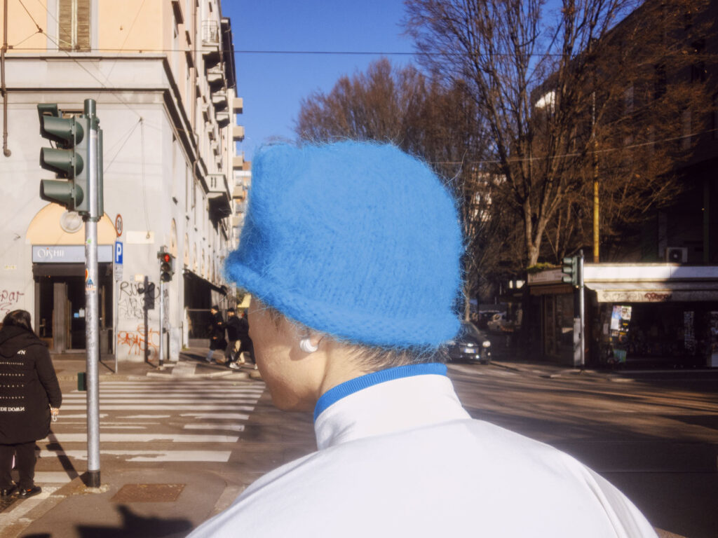 A street photography shot focusing on the back of a person wearing a bright blue fuzzy hat and a white top, standing at a city intersection.