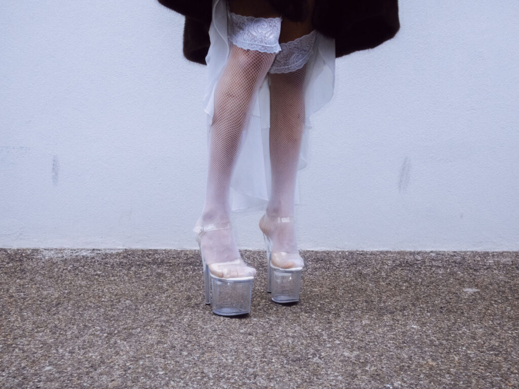 A close-up of a person's legs wearing white fishnet stockings, lace garters, and clear platform heels.