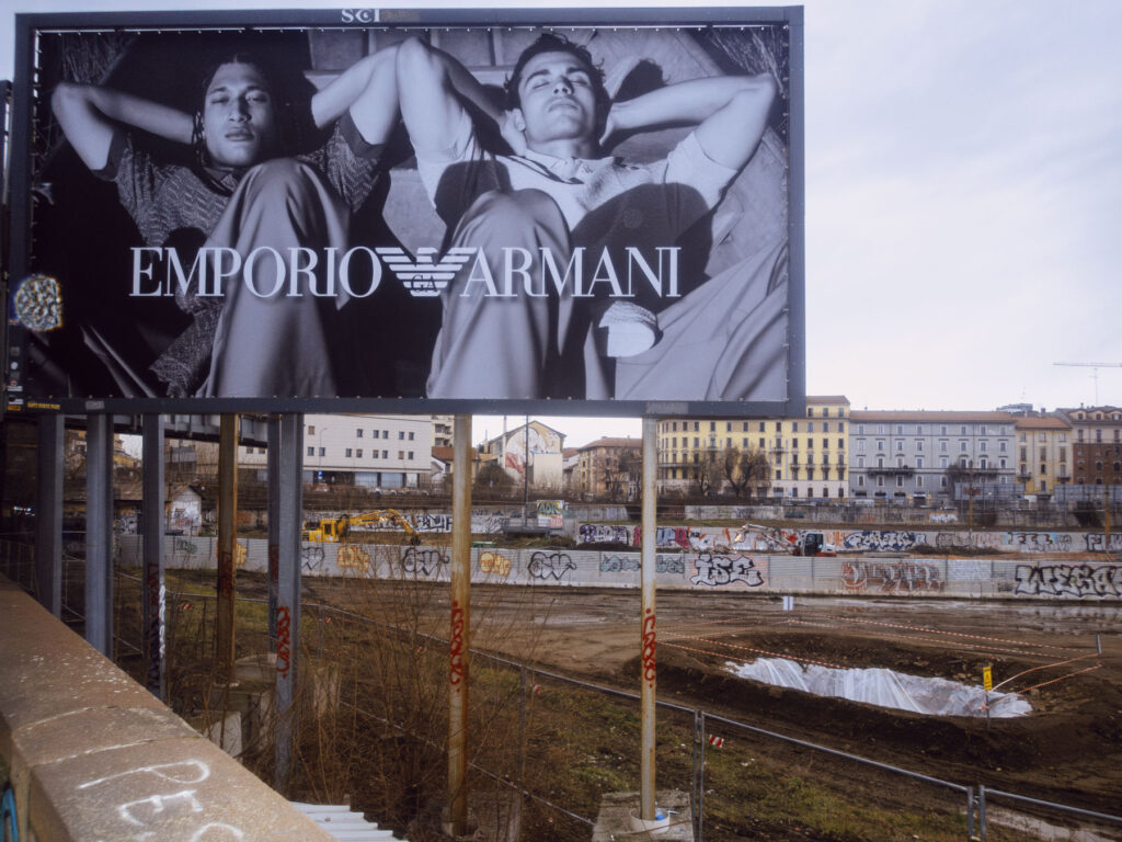 A street photography shot contrasting a luxury Emporio Armani billboard featuring reclining models with a construction site below, where a large pit covered in plastic mirrors the models' relaxed posture. Graffiti and urban decay surround the scene, highlighting the clash between elegance and reality.