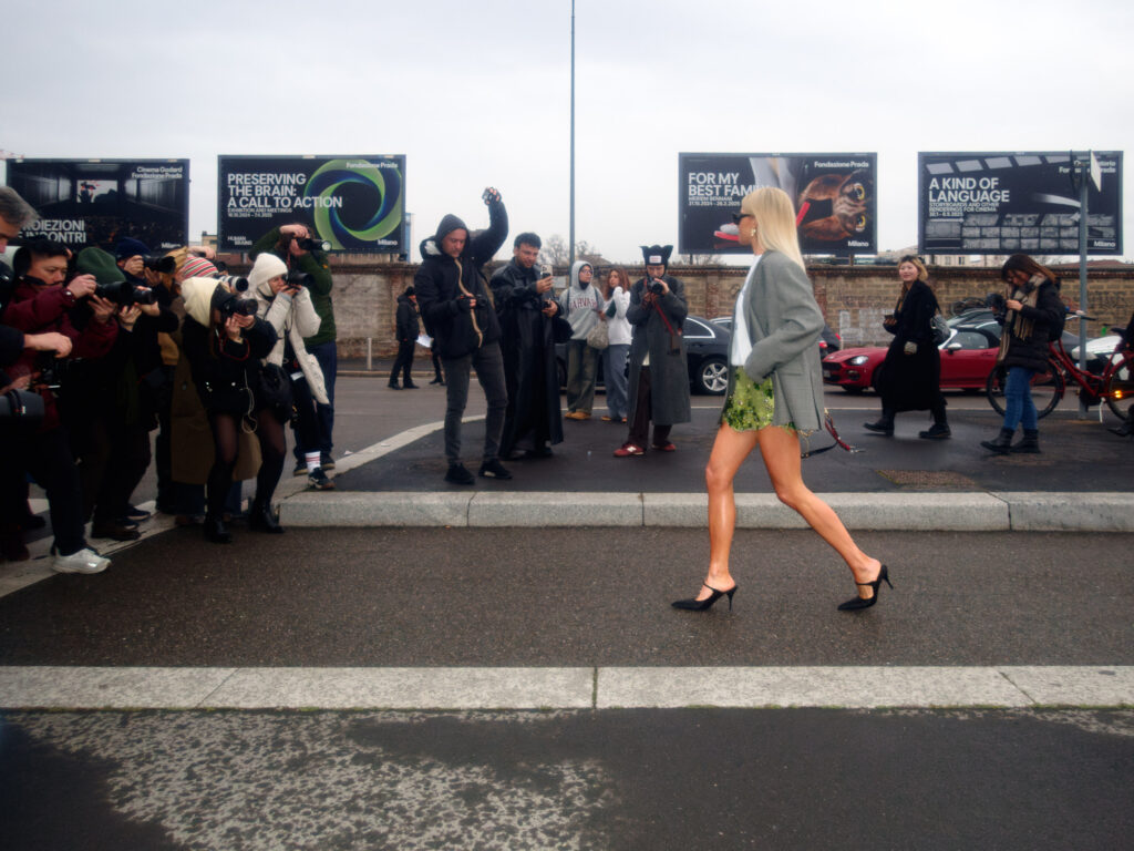 A fashion-forward woman with platinum blonde hair confidently strides across the street in heels and a blazer, while a crowd of photographers captures the moment. Billboards and onlookers form the urban backdrop.