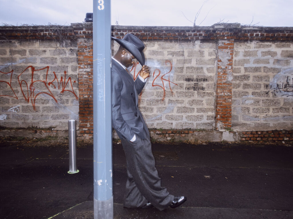 A stylish street photography portrait of a man leaning against a pole on a gritty urban street. He wears a sharp grey suit, a wide-brimmed hat, and polished black shoes, exuding a classic and confident style. The background features a weathered brick and stone wall adorned with red graffiti, contrasting with the subject's polished appearance. The overcast sky and muted tones of the setting add to the raw, cinematic atmosphere of the scene.