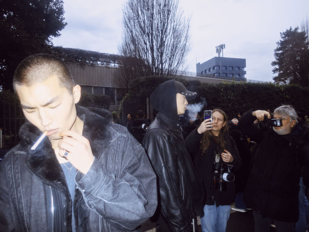 A scene capturing a group of people in an urban setting. In the foreground, a man with a buzz cut smokes a cigarette, wearing a fur-lined denim jacket. Nearby, another man in a black leather jacket and cap exhales smoke as a another one photographs him with her phone. A photographer with a flash camera captures the moment from the side, while others linger in the background.