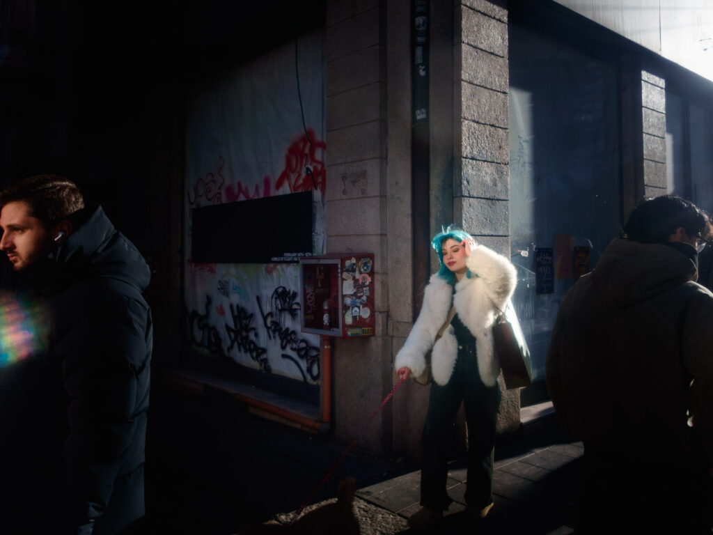 A street scene with dramatic lighting: a woman with blue hair and a white fur coat is illuminated by sunlight, standing against a graffiti-covered wall while holding a red leash. Shadows and dark-clad pedestrians frame the composition.