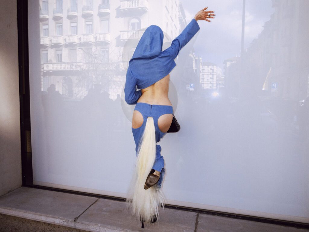 A person is performing a handstand wearing a blue hoodie and denim shorts, with a long ponytail extension attached at the back of the shorts, cascading downwards, creating a unique and playful visual effect.