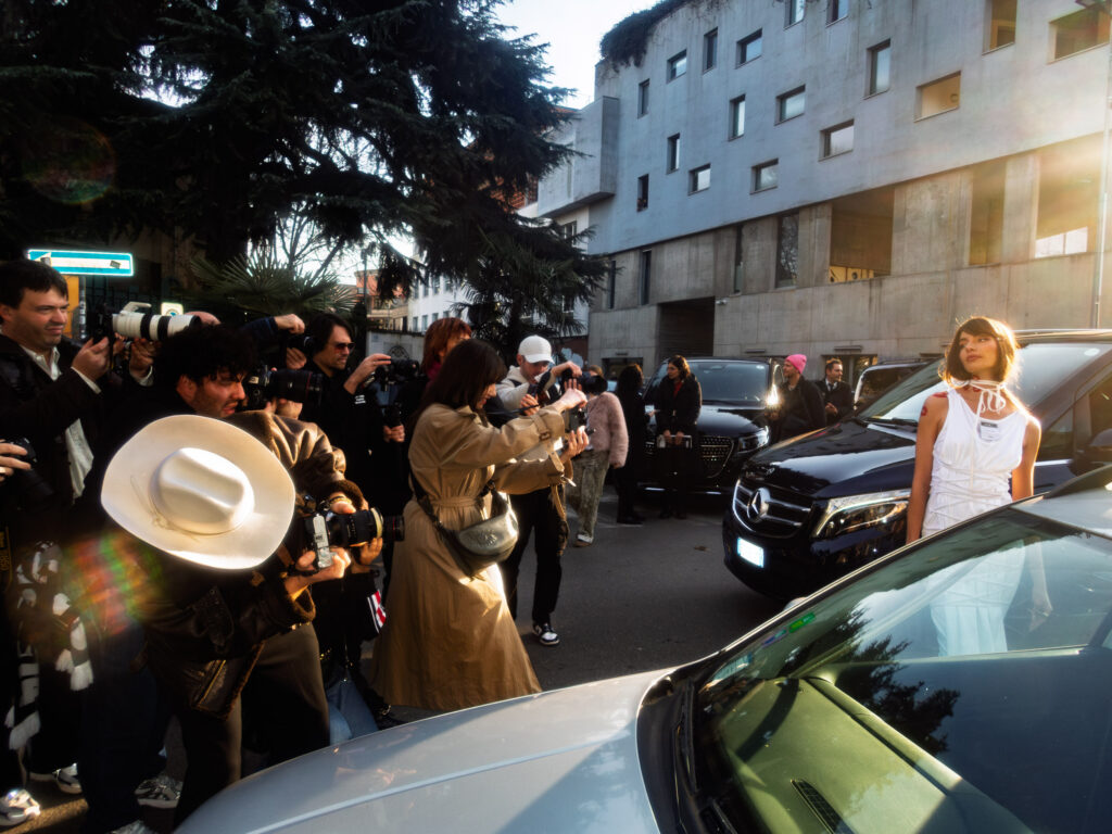 A bustling outdoor scene during Milan Fashion Week, where a group of photographers in diverse outfits crowd around a woman in a white, avant-garde dress standing near parked cars. She looks poised and confident while the cameras capture her from multiple angles under the warm afternoon light.