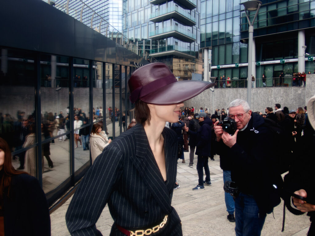 A stylish model wearing a wide-brimmed burgundy hat and a pinstriped blazer stands amid a bustling crowd during Milan Fashion Week. A photographer in the foreground captures her from a close angle, while modern glass buildings and onlookers reflect in the windows behind them.