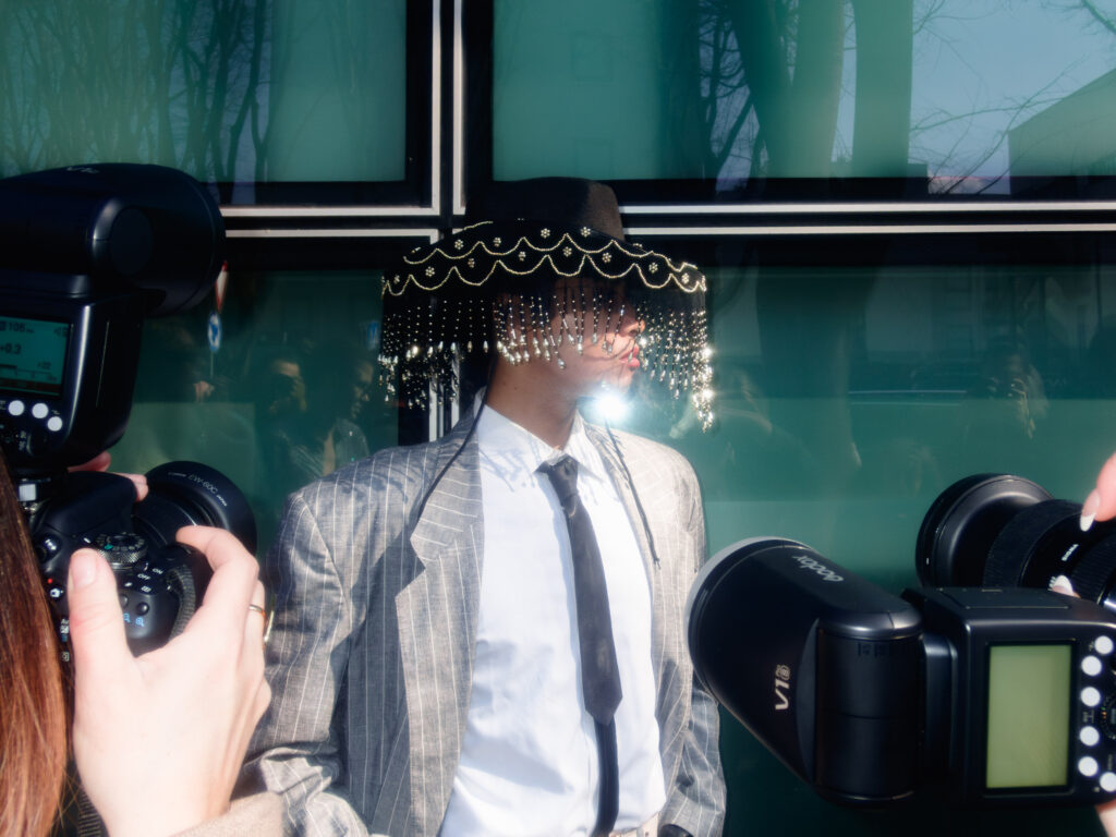 A fashion-forward attendee, wearing a tailored pinstriped suit and a dramatic black fringe hat adorned with shimmering beads, is photographed up-close by multiple cameras during Milan Fashion Week. The hat’s dangling ornaments partially obscure the wearer’s face, reflecting flashes and adding a dazzling effect against a green glass backdrop