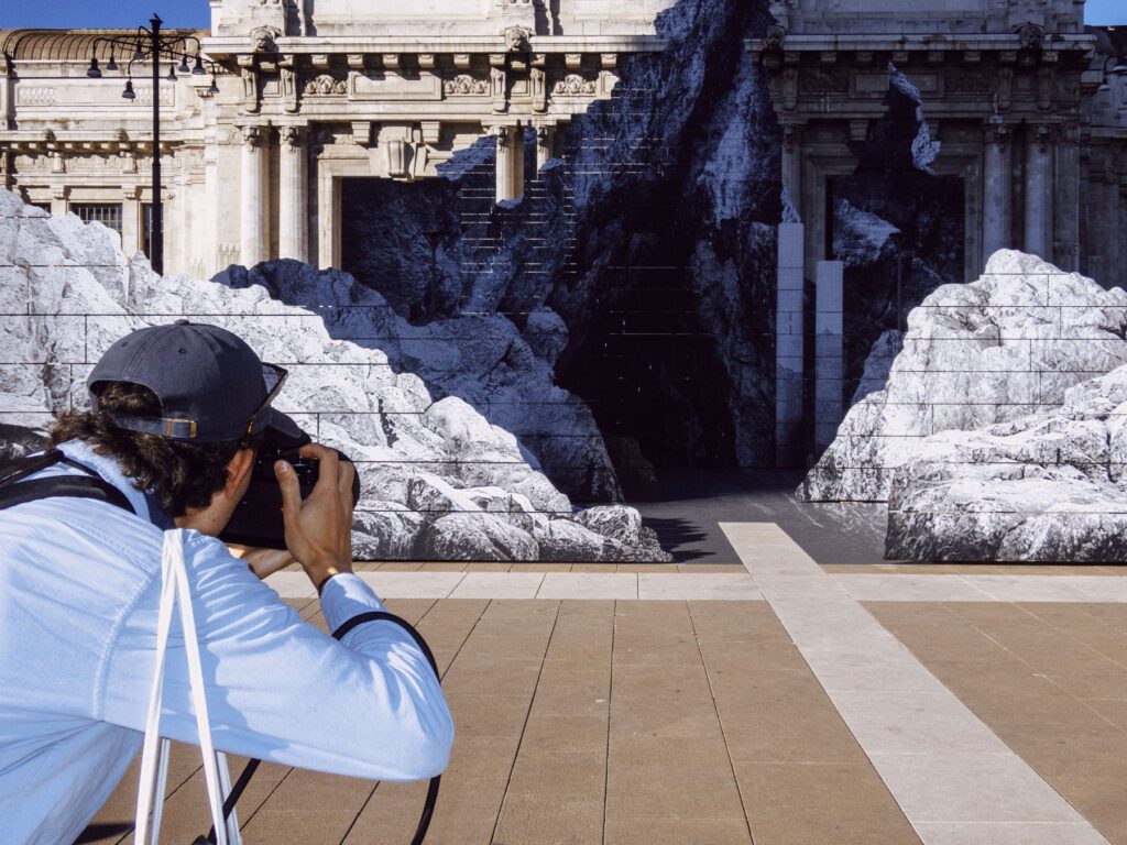In this photo, a photographer is captured in the act of taking a picture of a striking art installation. The installation features realistic mountainous shapes that contrast sharply with the classical architecture in the background, creating an intriguing visual blend. The photographer, dressed casually with a cap and a light jacket, is focused on his subject, illustrating the act of documenting the interplay of art, history, and public space.