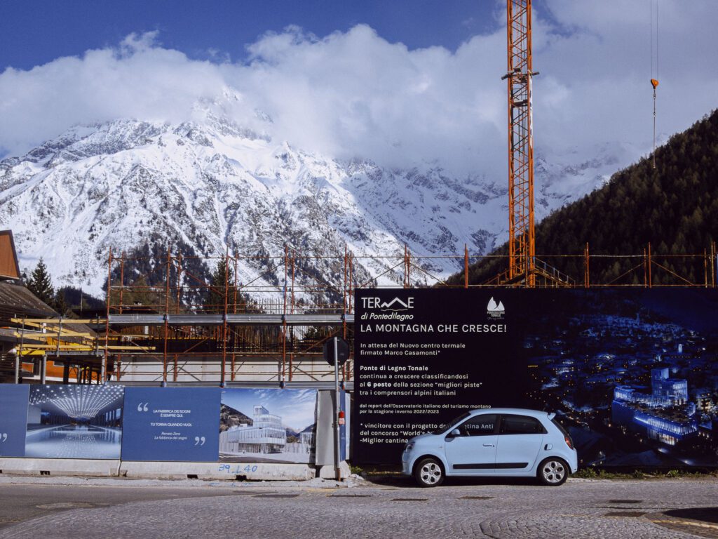 This image captures a striking contrast between nature and development. In the foreground, there's a construction site with scaffolding and a large crane. Promotional banners around the site announce a new thermal center, with renderings of the future building. A small light blue car is parked in front, accentuating the scale of the construction. Behind this scene of progress, the majestic, snow-capped mountains stand tall, underscoring the juxtaposition of human activity against the backdrop of the natural world.