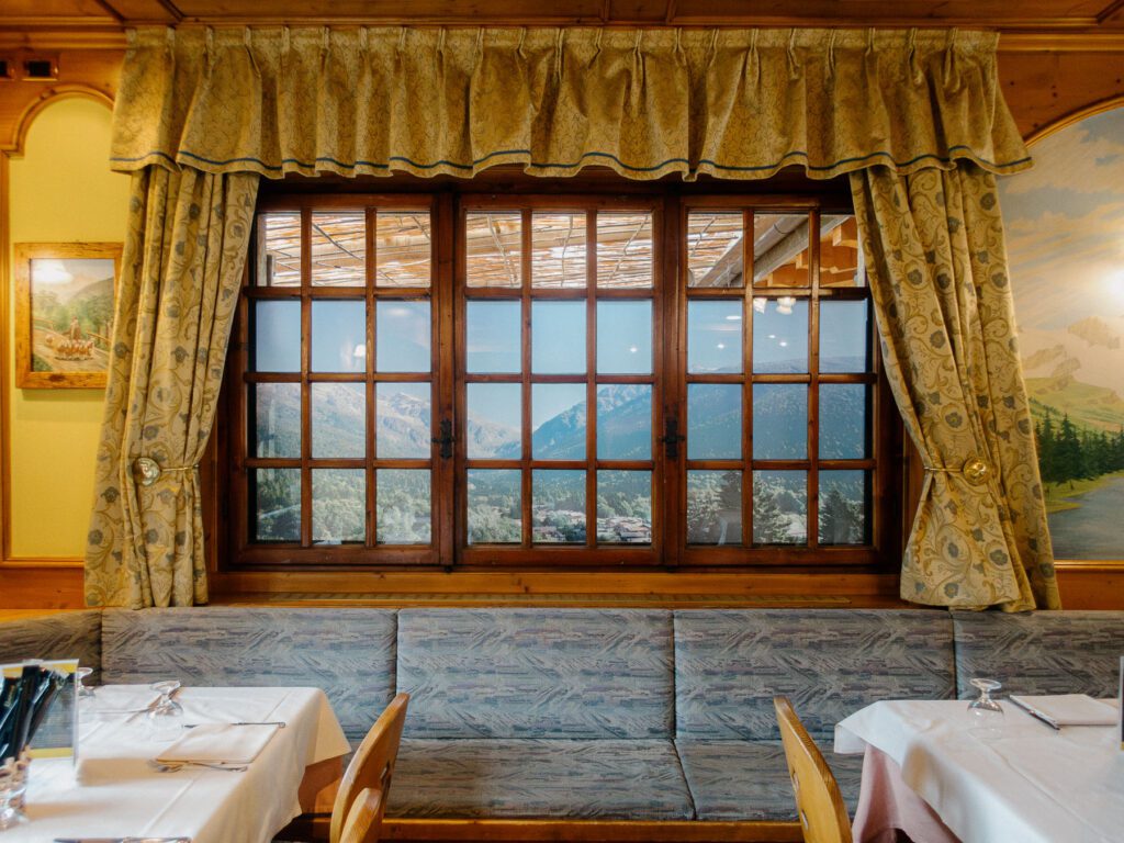 Inside a mountain-themed restaurant, a large window with patterned curtains reveals not the outdoors, but a wall-sized poster simulating a mountain view. The dining area is inviting, with white tablecloths on the tables and benches along the wall, all contributing to a charming and rustic atmosphere that evokes the essence of a cabin retreat.