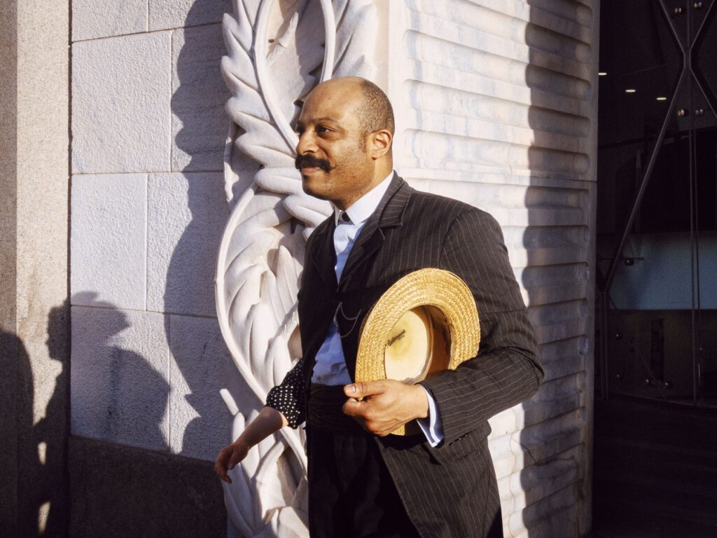 A man in a pinstripe suit holds a straw hat as he stands in front of an intricately carved stone wall. The scene is well-lit with natural sunlight, casting distinct shadows on the wall. The man's expression is calm and he appears to be mid-step. A woman's arm, visible in the background, suggests she is walking with him.