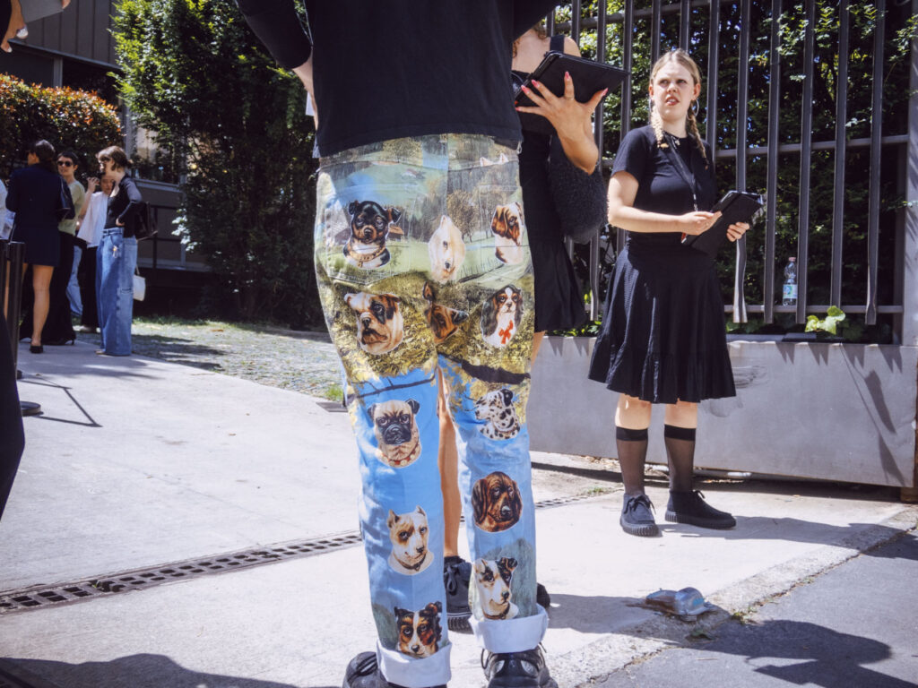 A street photography scene depicting a man wearing pants with vibrant dog portraits printed on them, standing in the foreground. He is accompanied by a woman dressed in a black skirt and top, holding a tablet, looking to her side with an expressive face.