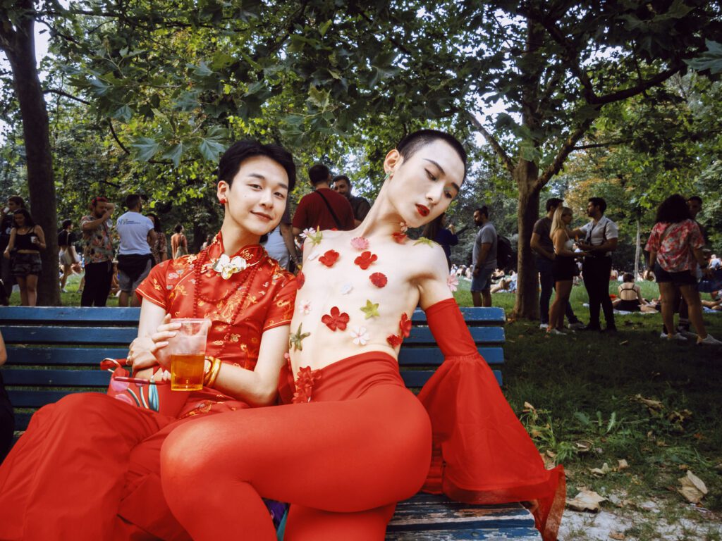 Two individuals dressed in striking red outfits sit on a park bench during Milano Pride 2024. The person on the right poses confidently, adorned with floral decorations on their upper body. The background features other attendees enjoying the event amidst the greenery of the park.