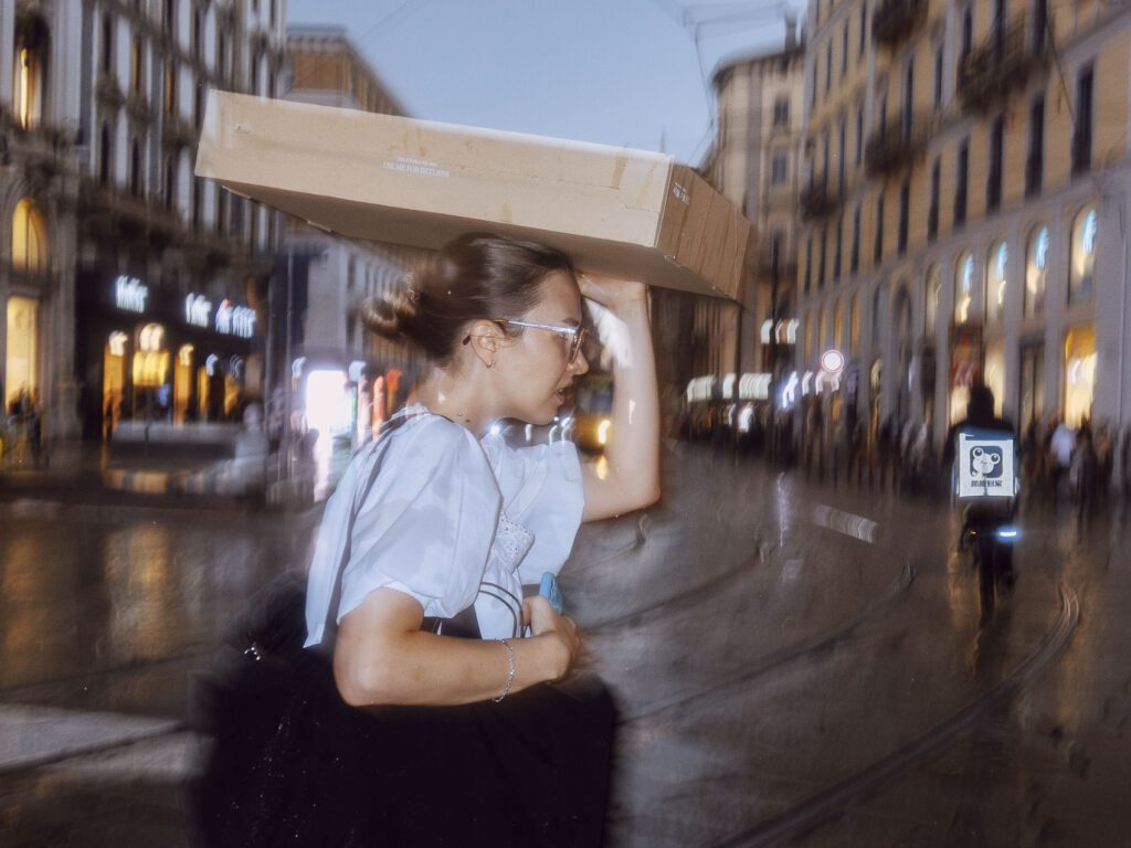 A woman carrying a cardboard box on her head walks through a busy, rain-soaked urban street at dusk, with blurred lights and buildings in the background.