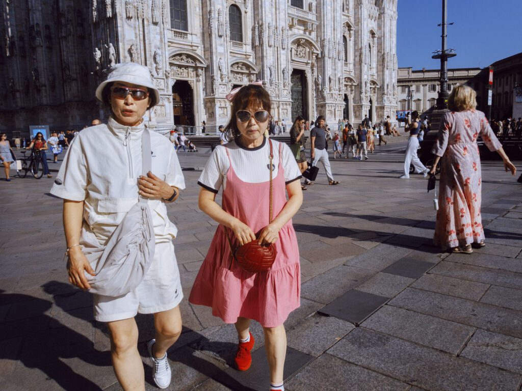 Two people walking in front of the Milan Cathedral (Duomo di Milano) on a sunny day. The person on the left wears a white outfit with a bucket hat and sunglasses, while the person on the right wears a pink dress over a white t-shirt, also with sunglasses. The background features the detailed facade of the cathedral and various people walking and engaging in activities on the piazza.