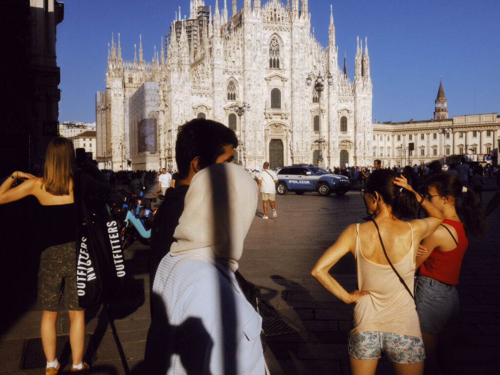 A bustling scene in front of the Milan Cathedral (Duomo di Milano) in the late afternoon sun. The image features various people, including a woman in a black top with a large "Outfitters" shopping bag, a person with a white headscarf, and two women in summer attire standing with their backs to the camera. A police car is parked in the background, and the cathedral's stunning gothic architecture is illuminated by the sunlight.