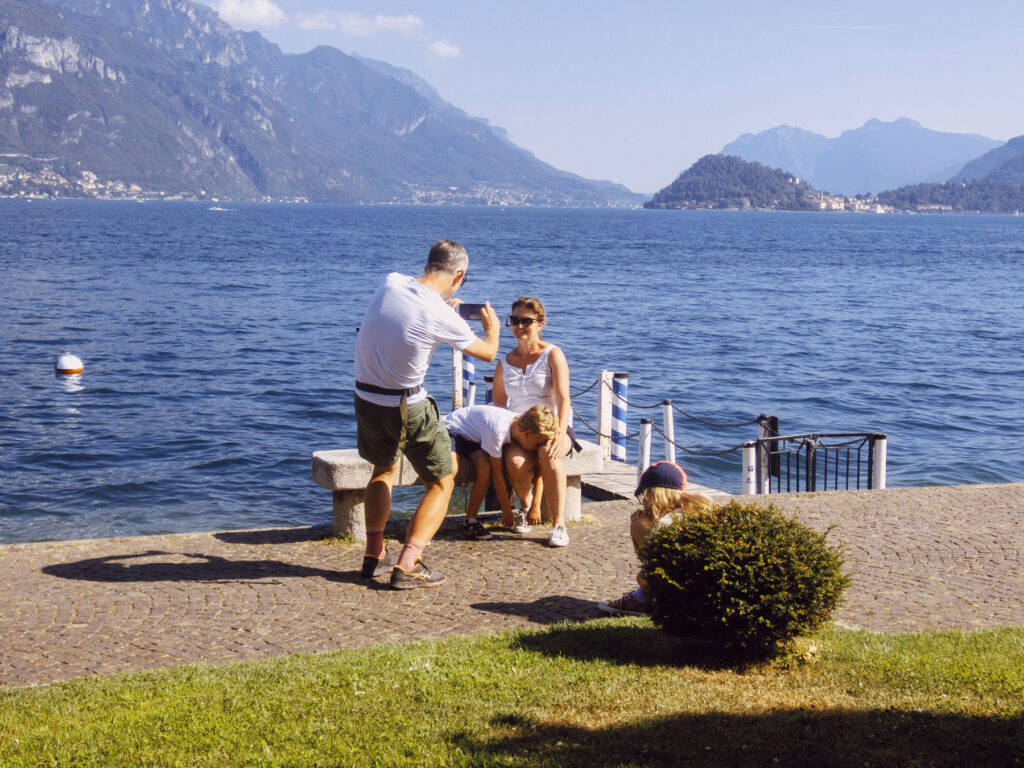 A serene lakeside scene featuring a family enjoying a sunny day. A man in shorts and a white t-shirt crouches with a smartphone, taking a photo of a woman sitting on a bench with her tired and exhausted young son resting on her legs. Nearby, a young girl wearing a hat sits on the ground. In the foreground, a small bush adds greenery, while the background showcases a stunning lake surrounded by majestic mountains under a clear blue sky.