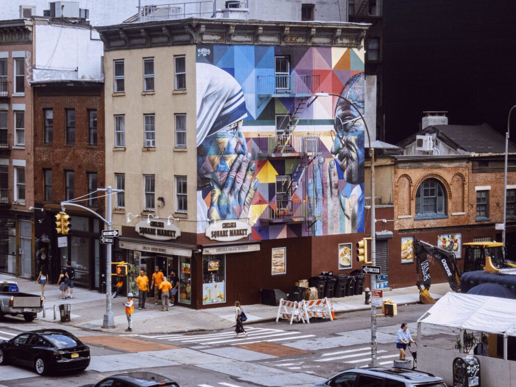 Urban street corner with a large, colorful mural on a building, depicting abstract geometric shapes and a photorealistic image of a person. Pedestrians walk by and cars drive on the road. Construction equipment and barriers are also visible