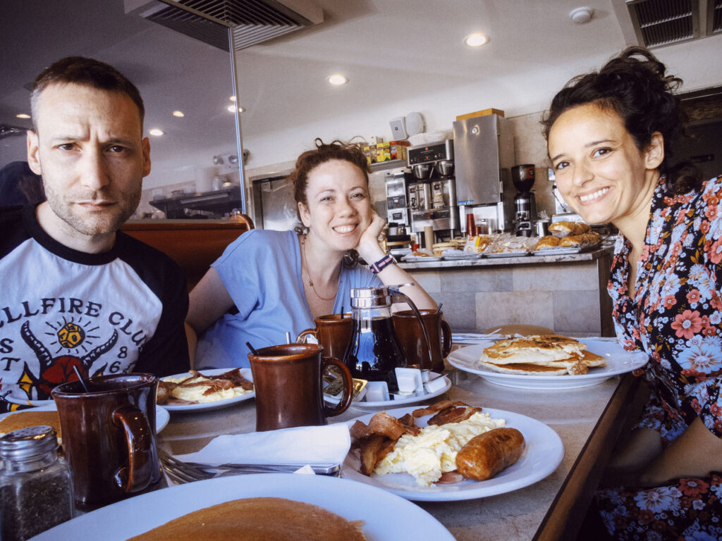 An image capturing three friends seated at a diner table, enjoying a hearty breakfast. The man on the left wears a graphic t-shirt and looks directly at the camera with a serious expression. The two women next to him, one in a light blue top and the other in a floral dress, smile warmly. Plates of pancakes, eggs, bacon, and sausage are on the table, along with mugs of coffee and syrup. The diner setting in the background features a counter with pastries and coffee machines, adding to the casual, vibrant atmosphere.