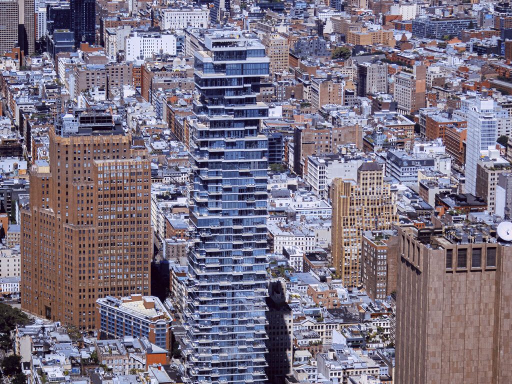 Aerial view of a dense cityscape with a variety of buildings. A distinctive building with a series of stepped terraces stands out in the center, surrounded by traditional high-rise buildings. The pattern of the city blocks is visible, with a mix of architectural styles and rooftop features.
