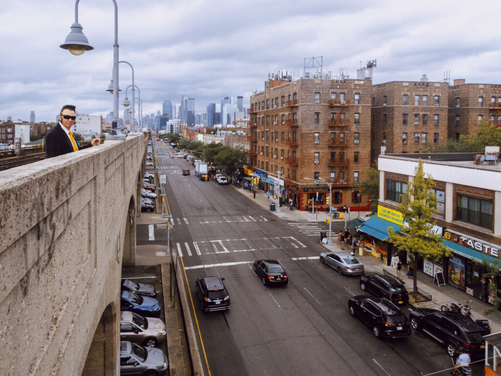 An urban scene featuring a wide street lined with brick buildings and small shops, including a farmer's market and a restaurant. Cars are driving along the street, with others parked on the sides. On the left, a man with a look reminiscent of Elvis Presley, dressed in a dark suit, sunglasses, and a yellow scarf, stands on an elevated concrete walkway, observing the scene below. In the background, the modern skyline of a city with tall skyscrapers is visible under a cloudy sky.