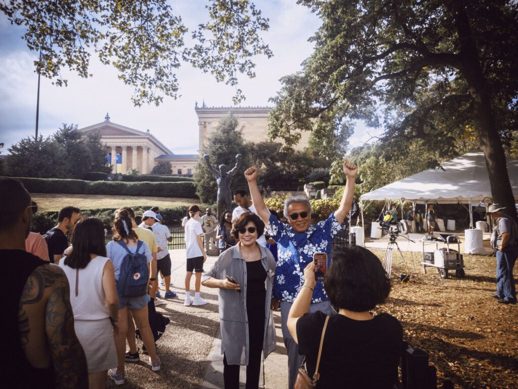 A group of people are gathered outdoors near a statue of a person with raised arms. In the foreground, an elderly man wearing sunglasses and a blue floral shirt is raising his arms in celebration, while a woman next to him is smiling. Another woman is taking their photo. In the background, a large building with columns, likely a museum, is visible, along with a tent and various equipment. Trees provide shade to the area.