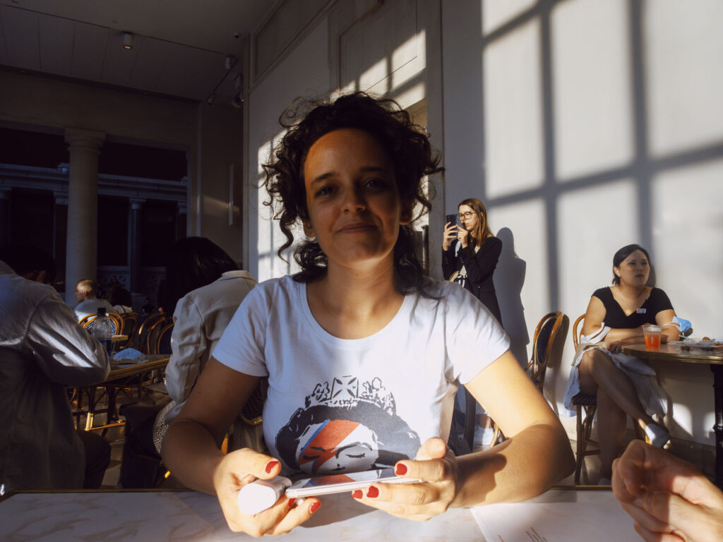 An image taken indoors, capturing a woman sitting at a sunlit table with soft shadows cast across the room. She wears a white t-shirt with a bold design, holding a phone in her hands, and smiles subtly toward the camera. Behind her, a mix of people are engaged in different activities: a woman photographing with her phone, another seated with a drink, and others immersed in conversation. The setting, with columns and large windows, blends natural light and architectural elegance, creating a layered composition that highlights everyday human interactions.