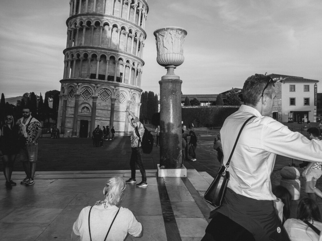 Tourists are taking playful photos in front of the Leaning Tower of Pisa during sunset. A man in the foreground pretends to hold up the tower with his hand, a common pose for pictures at this famous site. A couple poses for a photo, and other visitors are scattered around the square, enjoying the landmark and the warm glow of the setting sun.