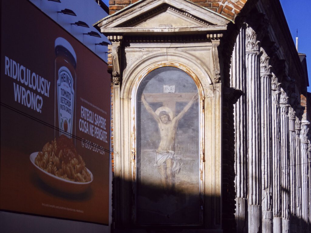 An image juxtaposing a religious fresco of Christ on the crucifix with a modern advertisement for Heinz Tomato Ketchup on pasta. The ad features a bold question, "Ridiculously wrong?" and a phone number with an invitation for feedback. This contrast highlights a stark clash of cultural imagery—sacred art versus commercial promotion—set against an architectural backdrop with classical column details. The scene captures a provocative and possibly controversial mix of traditional and contemporary elements.