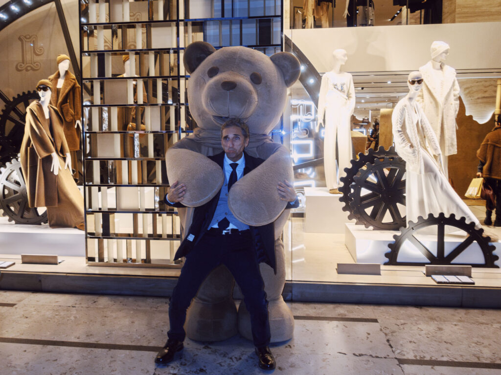 A man in a formal suit poses humorously in front of a large teddy bear statue, appearing to hold its oversized arms. The setting is a high-end fashion store window display featuring mannequins dressed in luxurious clothing, including tailored coats and elegant outfits. The display incorporates mechanical gears and reflective surfaces, adding a modern and industrial aesthetic to the backdrop.