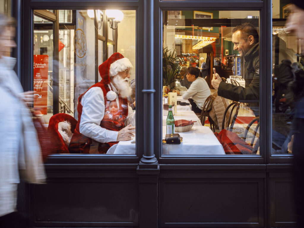 Scene capturing a candid moment inside a café. A man dressed as Santa Claus, in his red suit and hat, sits at a table engaging with patrons. Reflected in the café window, a child gazes at Santa with curiosity and wonder, adding a magical touch to the composition. On the right, a man smiles while taking a photo of Santa with his phone. Outside, blurred figures of passersby add movement.