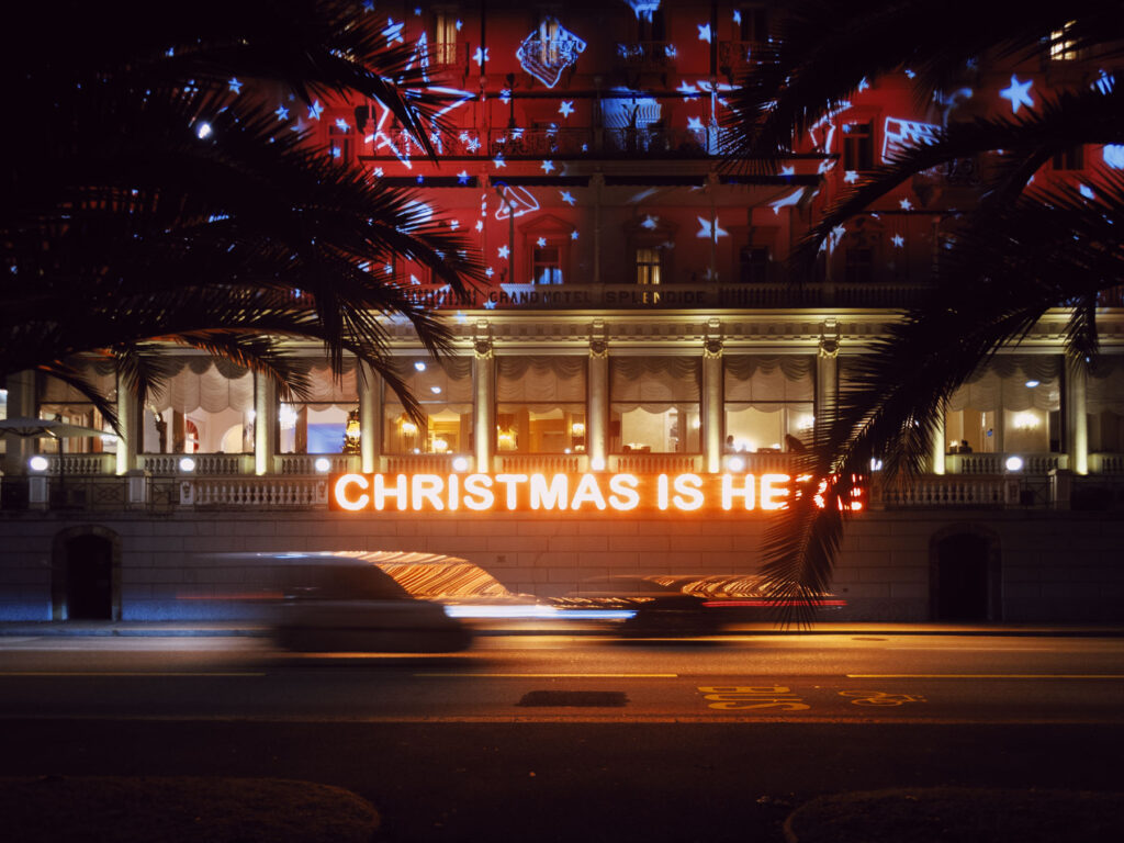 Scene featuring a grand, illuminated building at night with a bright red neon sign reading 'CHRISTMAS IS HERE.' The façade is decorated with projected stars and festive lights, creating a holiday atmosphere. Palm tree silhouettes frame the image, while a blurred car passes in the foreground, emphasizing motion. The combination of light, architecture, and movement conveys the energy and joy of the holiday season in an urban setting.