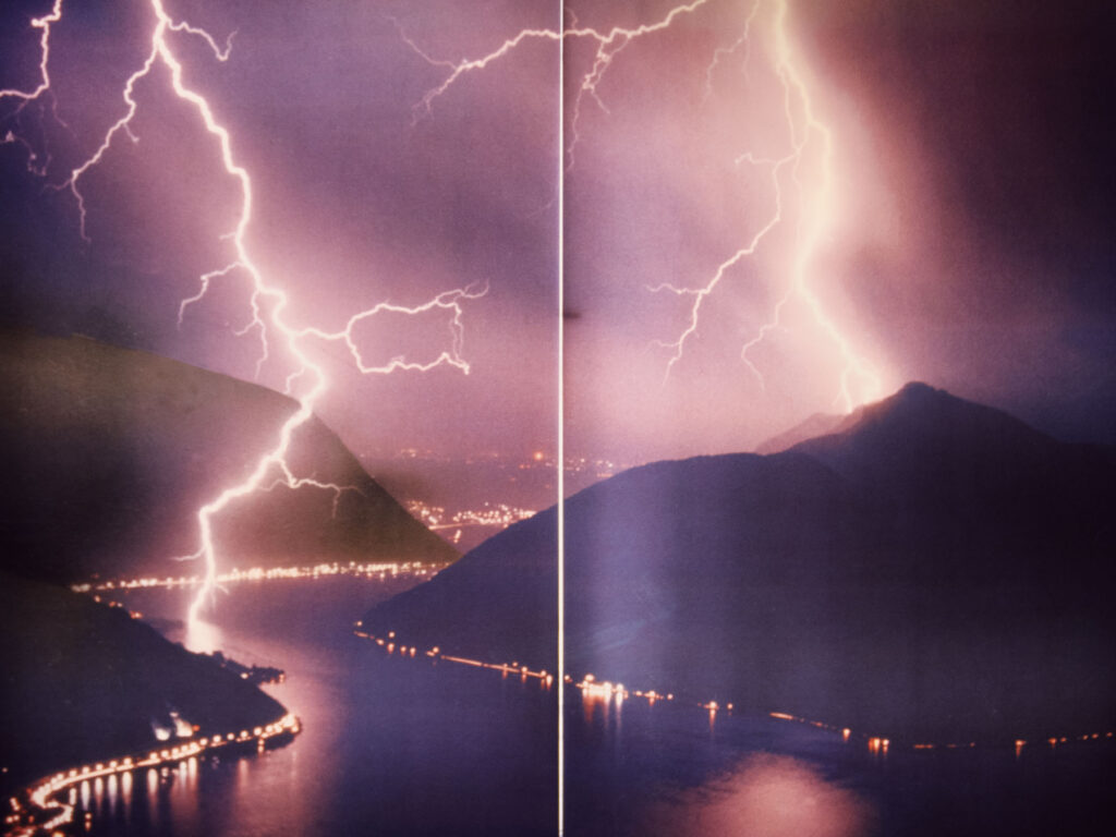 A dramatic nighttime image of two powerful lightning bolts striking over a mountainous landscape, captured on a large printed poster. The vertical line in the center reveals the fold or seam of the poster, indicating it as a physical print. The scene is filled with vibrant purple and pink hues from the lightning, which reflect off the calm waters of a lake below. The shoreline is dotted with glowing lights from towns and roads, blending the raw power of nature with the human presence in the serene landscape.