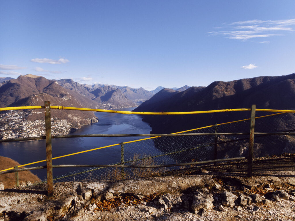 A scenic landscape photograph capturing a tranquil lake surrounded by towering mountains under a clear blue sky. In the foreground, a weathered yellow railing with signs of rust and a green wire fence frame the view, adding a rugged, textured contrast to the serene natural beauty. Small towns and villages dot the shoreline in the distance, nestled against the slopes, while sunlight highlights the contours of the hills and calm water.