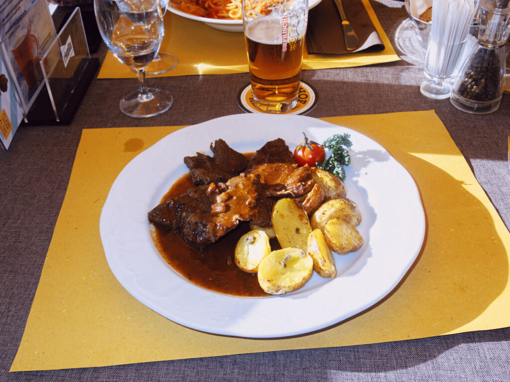 A close-up shot of a plate of food on a table set with yellow placemats. The dish features braised meat in a rich sauce, accompanied by golden roasted potatoes, a cherry tomato, and a small sprig of parsley. A glass of beer, a wine glass, and a plate of spaghetti are visible in the background, suggesting a dining setting. The sunlight casts soft shadows, adding warmth to the composition and highlighting the textures of the meal.