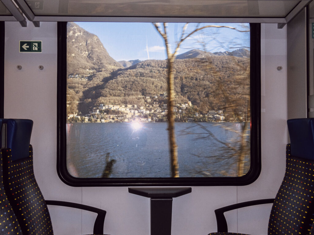 A scenic view of a lake and hillside town framed through the window of a moving train. The landscape features calm blue waters, dense wooded hills, and clusters of buildings along the shoreline under clear skies. A tree trunk appears blurred in the foreground, emphasizing the motion of the train. Inside the train, two empty seats with dark fabric and yellow dotted patterns flank a small table beneath the window, creating a contrast between the dynamic outside view and the still interior.