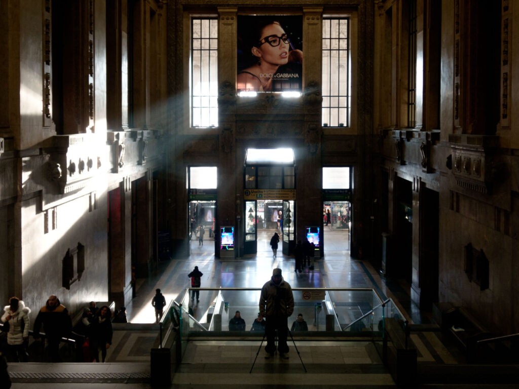 A dramatic interior shot of a grand train station hall, illuminated by beams of sunlight streaming through large windows. The architectural details of the high walls and ornate moldings create a sense of grandeur. In the foreground, a person stands still at the top of an escalator, silhouetted against the light. In the background, the bustling activity of travelers is visible, along with a large Dolce & Gabbana advertisement prominently displayed above the main entrance, adding a contemporary touch to the historic space.