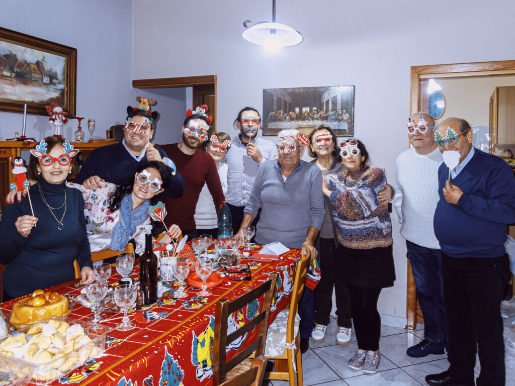 A cheerful family gathering during the Christmas season, with everyone wearing festive novelty glasses and accessories, including antlers and Santa hats. They stand around a dining table covered with a vibrant red tablecloth featuring Christmas motifs, set with desserts and glasses. In the background, a framed painting of The Last Supper hangs on the wall, adding a classic and symbolic touch to the scene.