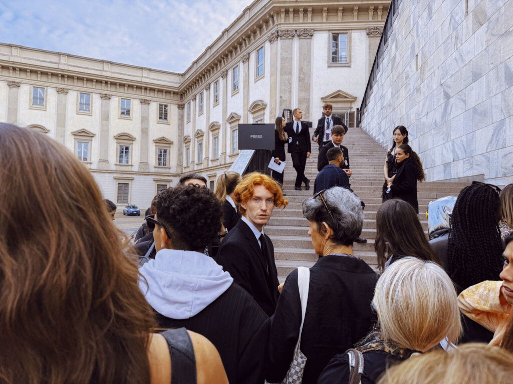 A candid street photography shot capturing a crowd gathered near a staircase leading to an elegant historic building. Among the crowd, a young man with striking red hair and a black suit stands out, looking directly at the camera with an intense gaze. The scene includes a mix of people engaged in conversation, some ascending or descending the steps, while others linger in groups. The backdrop features classical architecture and a sign reading 'PRESS,' adding a sense of occasion to the moment. The composition highlights individuality within the collective urban environment.