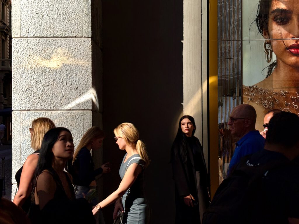 A street scene with people walking past a building. In the foreground, diverse pedestrians including women in casual attire and a person in a black robe. To the right, a large fashion advertisement shows part of a woman's face. Sunlight casts strong shadows on the textured white building wall.