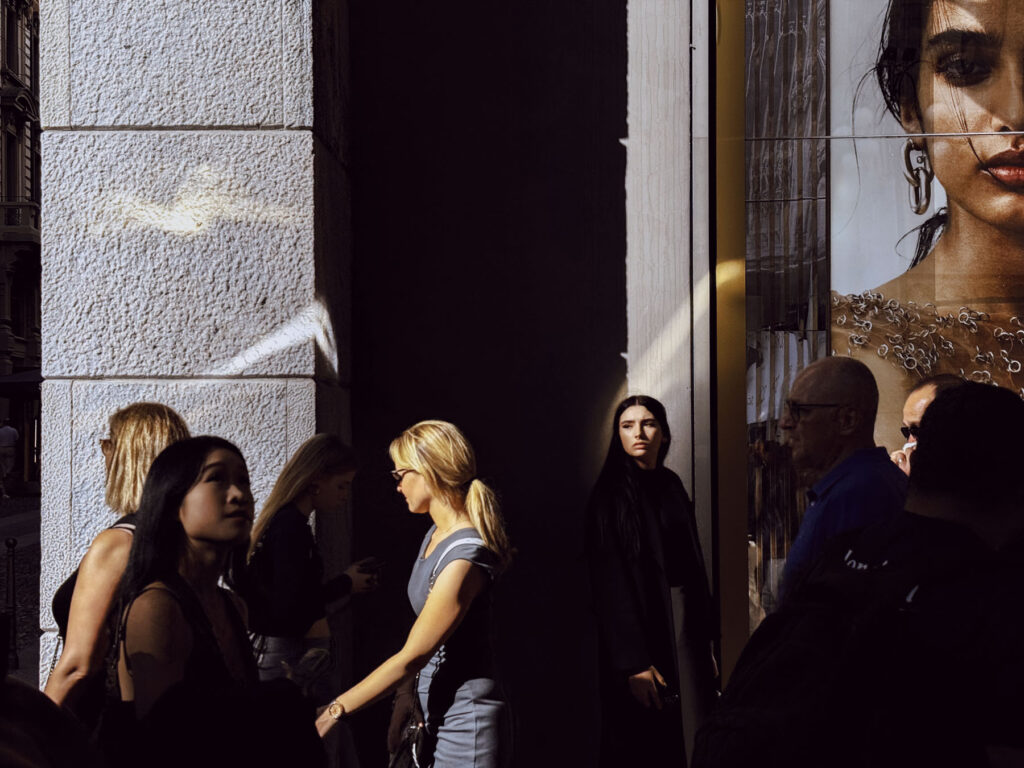 A scene capturing a group of people walking past a building with stark contrasts of light and shadow. The light highlights a young woman in black standing in the shadowed area, her gaze intense and reflective, creating a striking focal point. To the right, a large advertisement featuring a close-up of a model's face adds depth and contrast. The interplay of light and dark emphasizes the movement and emotions of the urban crowd, blending anonymity with individuality in a bustling city atmosphere.