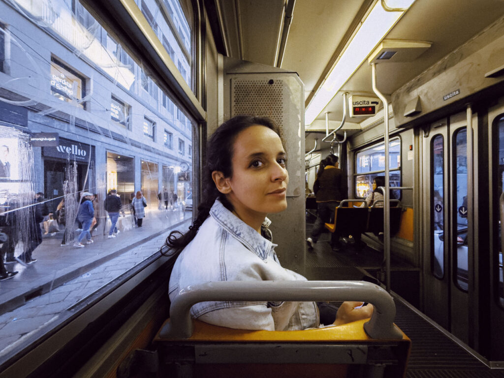 A woman sitting on a tram in a city, looking back towards the camera with a calm expression. The view outside the window shows a busy street with people walking by shops. The interior of the tram is modest, with a few other passengers seated in the background.