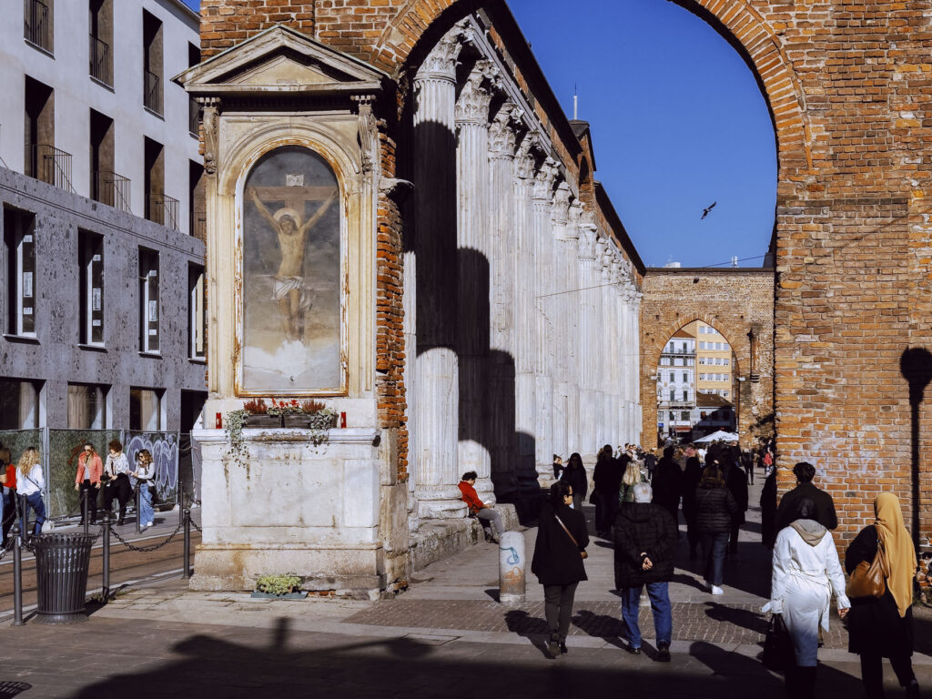 Historic Italian street scene with a religious shrine showing crucified Christ, framed by brick arches and marble columns. Pedestrians walk beneath the arches while a blue sky peeks through.