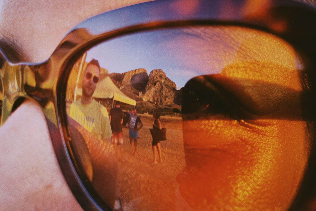 A close-up view of sunglasses reflecting a beach scene. The reflection shows a person wearing a yellow shirt standing on a sandy beach with other beachgoers nearby. In the background, rocky cliffs and a yellow tent or structure are visible. The sunglasses lens has a warm, golden tint, creating a summery, nostalgic atmosphere. The image has a vintage or film photography aesthetic with some light leaks or color distortions visible around the edges of the frame.