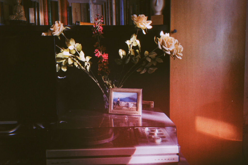 A dimly lit interior scene featuring a vase of flowers on top of what appears to be a record player or turntable. The flowers include light-colored roses and some red blooms. Next to the vase is a small framed photograph showing a person near water with mountains in the background. Behind the turntable is a bookshelf filled with books. The image has a warm, vintage aesthetic with reddish tones and film-like quality, suggesting it was taken with an analog camera or has a retro filter applied.