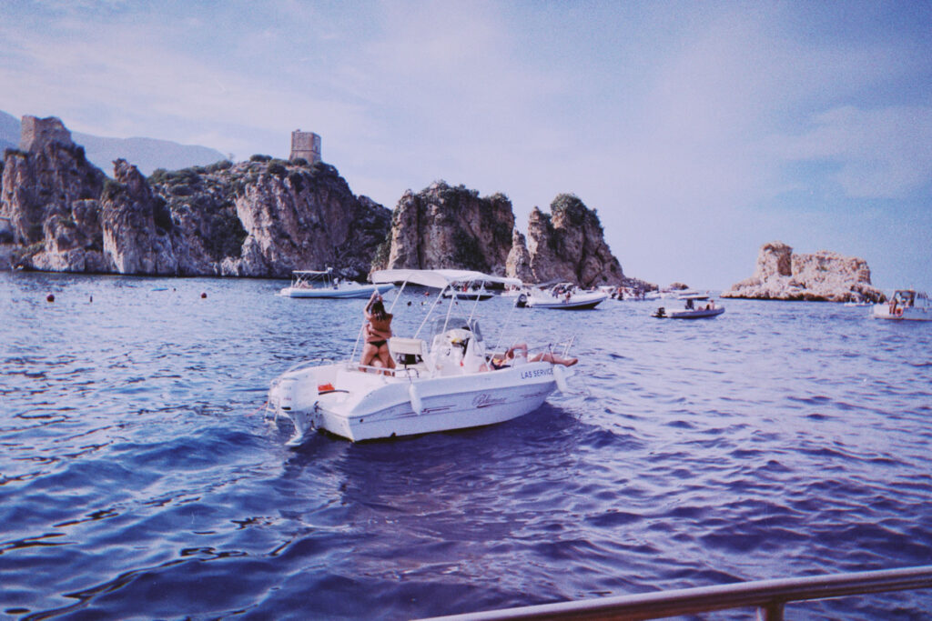 A scenic coastal view with rugged rocky cliffs and a tower-like structure on a hilltop. In the foreground, several small boats are anchored in calm blue waters. The main focus is on a white motorboat in the center, where a person in a swimsuit is standing. Other boats and people can be seen in the distance, enjoying the Mediterranean-like setting under a clear sky.