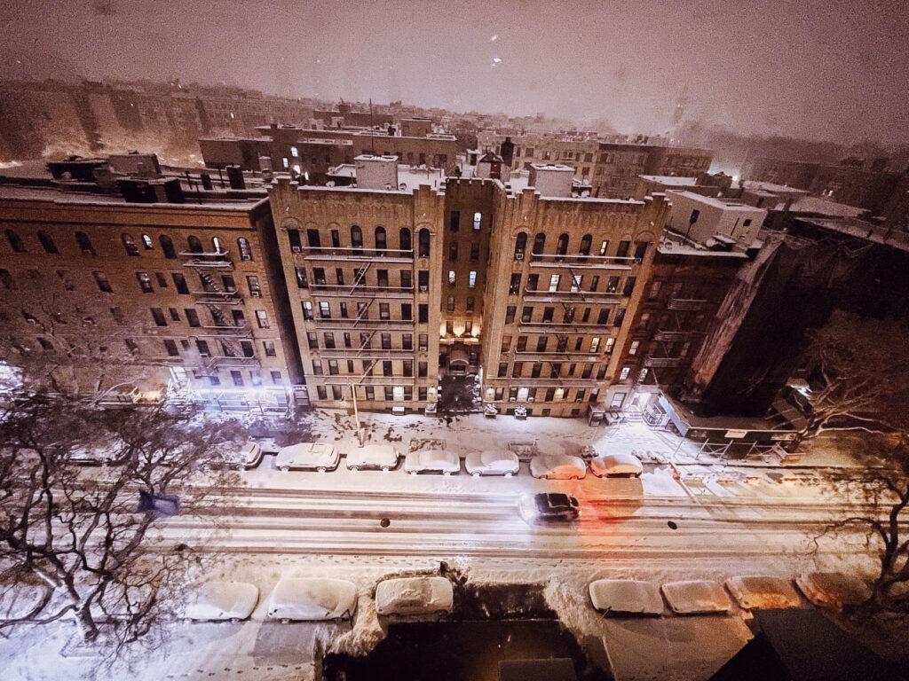 An image capturing a snowy urban scene at night, viewed from above. The focus is on a row of historic apartment buildings with intricate architectural details, framed by snow-covered streets and parked cars. Warm light spills from windows, contrasting with the cold, snowy atmosphere. A single car drives along the street, its headlights creating streaks of light on the snow. The cityscape fades into a hazy background, where the snow continues to fall, creating an atmospheric winter scene.