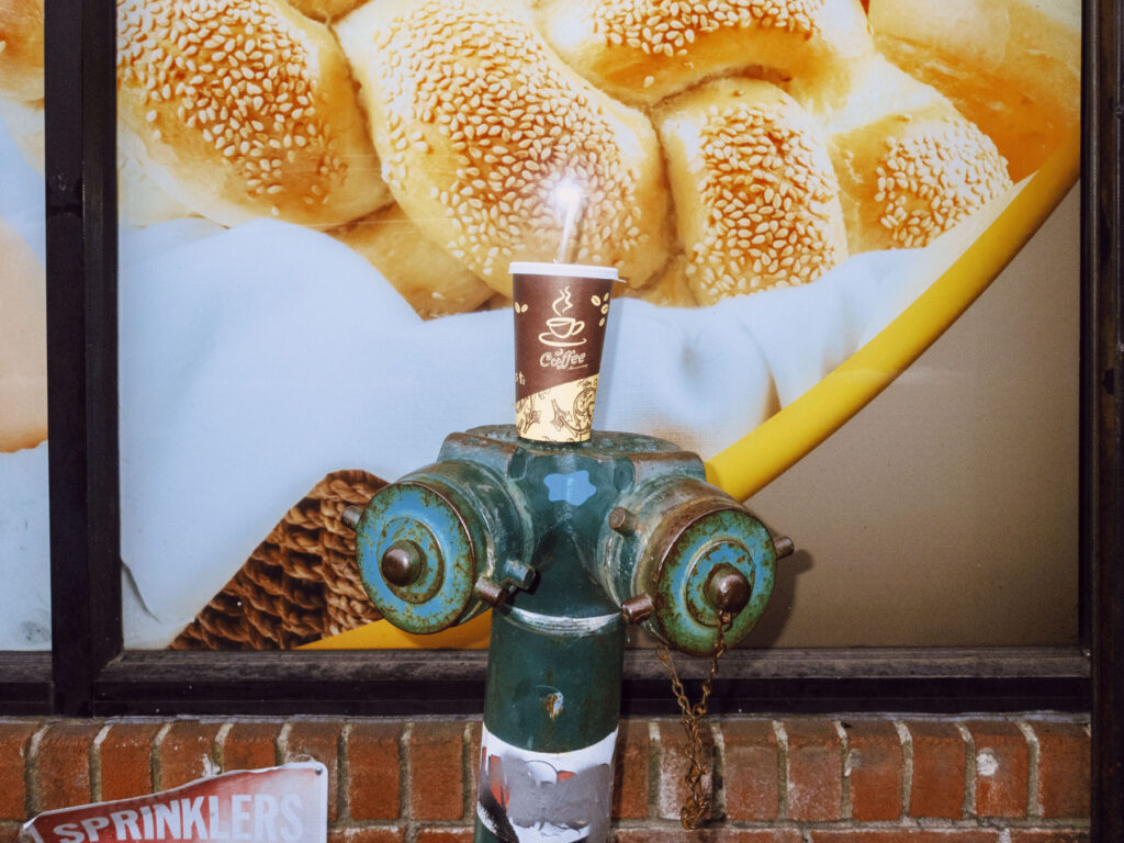 A quirky street photography shot of a green fire hydrant with a disposable coffee cup balanced on top. The backdrop features a large bakery advertisement with close-up images of sesame-covered bread rolls, creating an unexpected contrast between the urban and commercial elements. A brick wall and a partially visible 'SPRINKLERS' sign add texture to the composition, emphasizing the everyday yet artistic details of the scene.