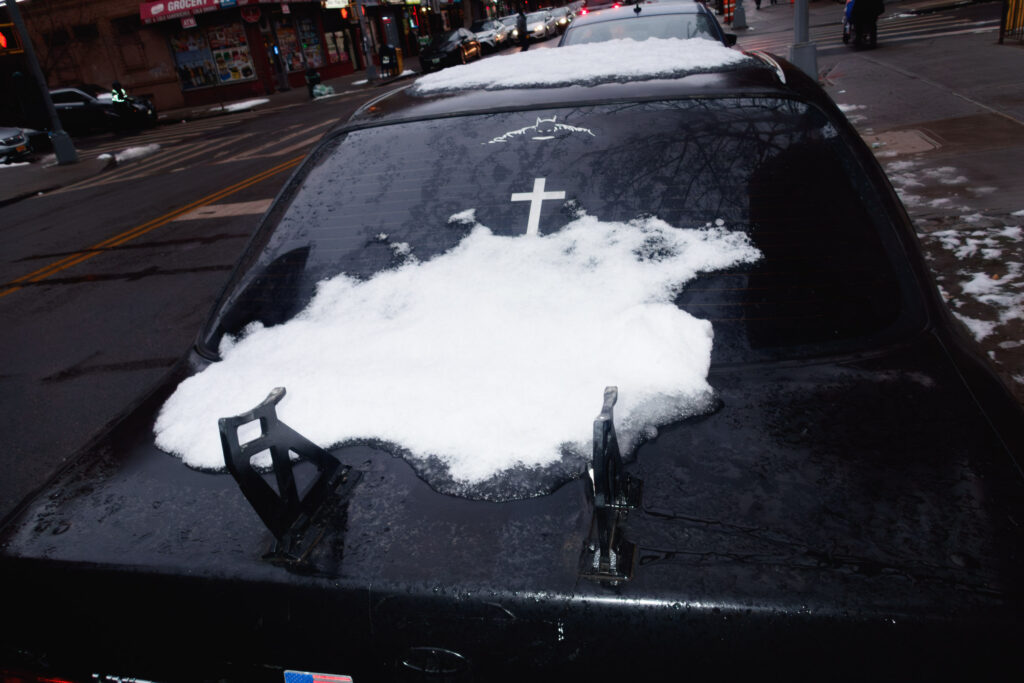 Photograph of a car's snow-covered rear windshield, with a small Batman symbol sticker and a hanging cross ornament inside it, parked alongside a city sidewalk. In the background, city storefronts and a sidewalk can be seen.